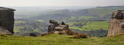 This landscape photograph features a panoramic view from Curbar Edge, an elevated escarpment in the Peak District, England. The image was captured in the late morning during autumn, as indicated by the subdued sunlight and the brown tones beginning to appear in the grassy foreground. The vantage point provides an extensive rural overview of the surrounding countryside in the Peak District National Park, with patchwork fields and wooded hills stretching into the distance. The village nestled below, typical of rural England, is visible with clusters of houses and winding roads. Rocky outcrops, characteristic of Curbar Edge, frame the scene, while a solitary figure sits on one of the rocks, emphasizing the scale of the landscape. This photograph highlights the natural beauty of this region in the United Kingdom, known for its dramatic viewpoints and open rural spaces.